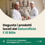 Due operatori del Salumificio F.lli Billo in divisa bianca mostrano un salume stagionato all'interno del laboratorio di produzione, con altri salumi appesi sullo sfondo. Evento di degustazione all'Interspar di Bassano del Grappa, C.C. Il Grifone.